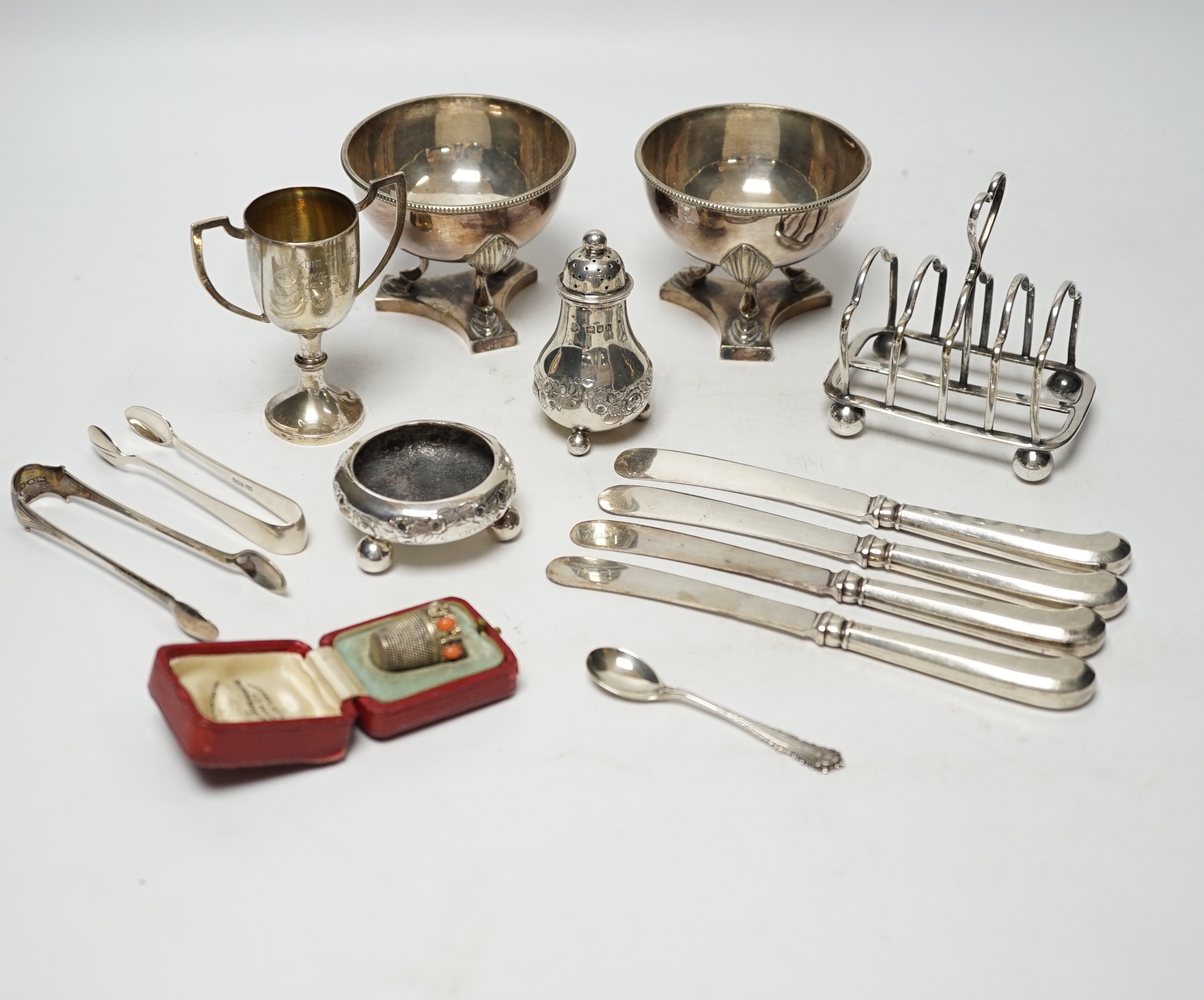 Twelve assorted silver condiments, including a set of four Edwardian bun salts, William Hutton & Sons, London, 1903, a set of six silver handled tea knives, a small silver trophy cup, silver napkin ring, cased silver thi